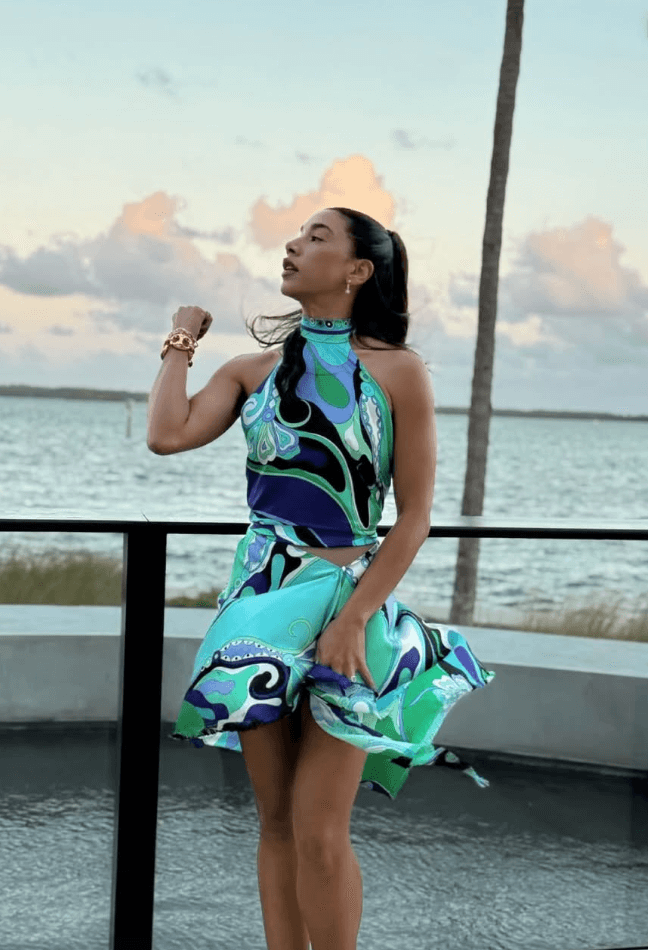 Carly Jordan Woman in a colorful, swirling-patterned blue and green dress stands by a waterfront railing, with palm tree and water visible in the background. Personal Stylist & Luxury Fashion Sourcer