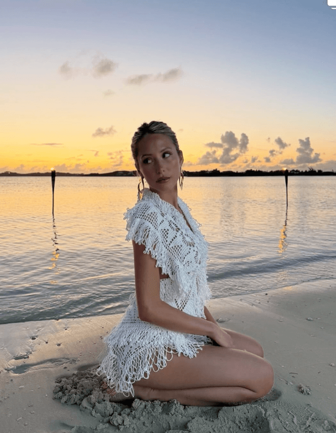 Carly Jordan Woman in a white crochet dress sits on sandy beach near water at sunset, with torches and clouds visible in the background. Personal Stylist & Luxury Fashion Sourcer