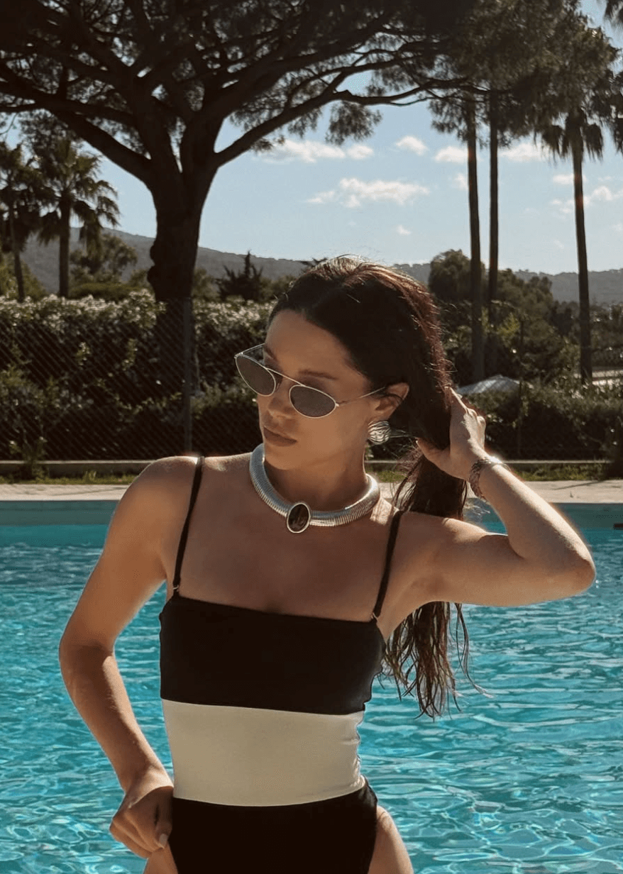 Carly Jordan Woman in a black and white swimsuit stands by a pool, wearing sunglasses and a large necklace, with trees and mountains in the background. Personal Stylist & Luxury Fashion Sourcer